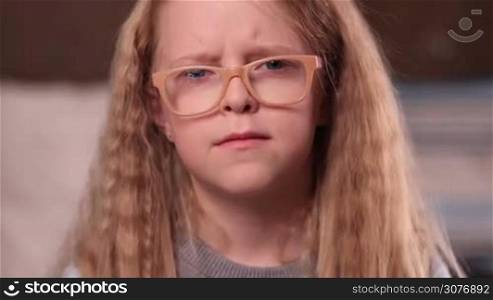 Closeup facial expression of upset little girl looking at camera. Sad little teen girl in glasses with amazing blonde long hair and blue eyes looking forward and ready to cry.