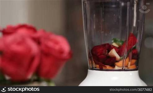 Closeup female adding chopped vegetables and fruits into blender shaker jug to prepare vegetable smoothie in household kitchen. Foreground blurry bouquet of red roses. Organic food and healthy eating lifestyle.