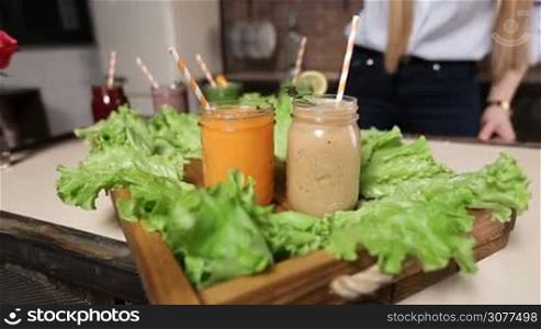 Closeup female hand serving wooden tray decorated with green salad with freshly blended fruit and vegetable smoothies of various colors and tastes in mason jars with straws and decors. Dieting, vegetarian, fitness, healthy lifestyle.