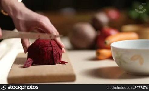Closeup female hands cutting beet into slices on wooden chopping board with kitchen knife. Side view. Woman slicing beet on cutting board and adding into ceramic bowl. Healthy vegetarian diet for weight loss and detox
