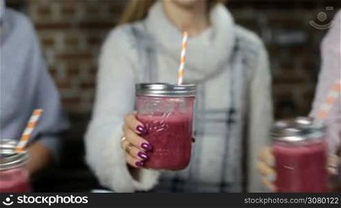 Closeup females hands toasting fresh blended berry smoothies in mason jars indoors. Midsection of women holding glass jars of detox strawberry smoothies and toasting over domestic background.