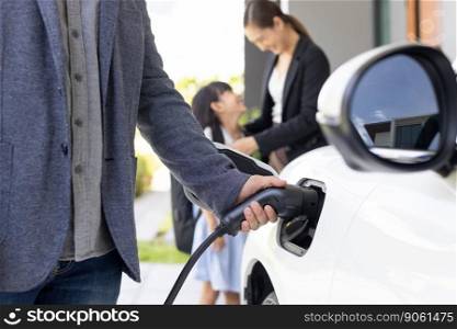 Closeup focus hand insert EV charger plug to electric vehicle at home charging station with blurred family in background. Progressive concept of green and clean energy to reduce CO2 emission by EV car. Closeup focus hand insert progressive EV charger plug to EV car at ho