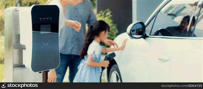 Closeup focus hand insert EV charger plug to electric vehicle at home charging station with blurred family in background. Progressive concept of green and clean energy to reduce CO2 emission by EV car. Closeup focus hand insert progressive EV charger plug to EV car at ho
