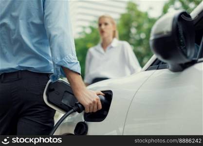 Closeup focus hand insert EV charger to electric vehicle at public charging point in car park with blur business people in backdrop, eco-friendly lifestyle by rechargeable car for progressive concept.. Closeup focus hand insert progressive EV charger with blurred background.