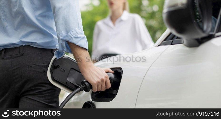 Closeup focus hand insert EV charger to electric vehicle at public charging point in car park with blur business people in backdrop, eco-friendly lifestyle by rechargeable car for progressive concept.. Closeup focus hand insert progressive EV charger with blurred background.