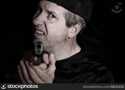 Closeup front view of mature man, looking forward and wearing baseball cap backwards, with gun in hand on dark background