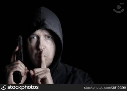 Closeup front view of mature man, looking forward and wearing hood, with weapon and index finger signaling to be silent on dark background