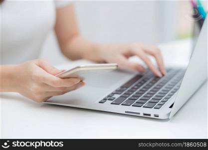 Closeup hand of young woman working laptop computer and reading smartphone on internet online on desk at home, freelance girl using phone with social media, business and communication concept.