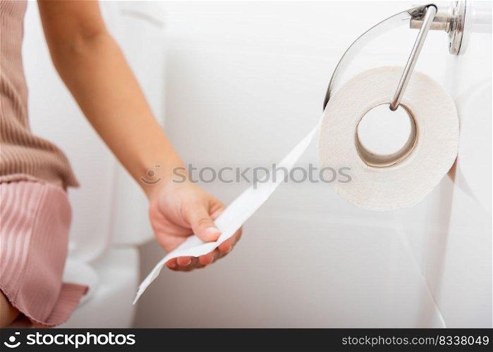 Closeup hand pulling toilet paper roll in holder for wipe, woman sitting on toilet she taking and tearing white tissue on wall to towel clean in bathroom, Healthcare concept
