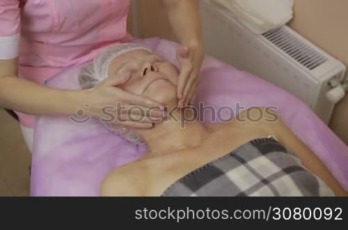 Closeup hands of beautican moisturizing senior woman&acute;s face with cream and massaging in beauty spa. Elderly woman getting face and neck massage in beauty salon. Cosmetician applying facial cream.
