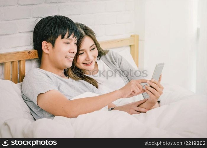 Closeup Happy Asian Lover or couple using technology smart phone on the bed in bed room at modern home, boyfriend pointing to the mobile phone, Lover and life style concept,