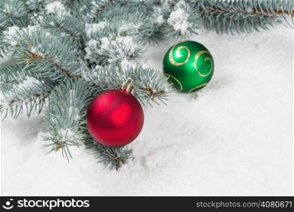 Closeup horizontal image of Christmas ornaments, red and green, hanging from a real Blue Spruce tree branch placed on snow