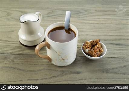 Closeup horizontal photo of a full cup of coffee, snacks and cream in pouring spout with aged wood underneath