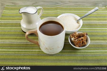 Closeup horizontal photo of a full cup of coffee with sugar and spoon in bowl, snacks and cream in pouring spout with green striped textured cloth underneath