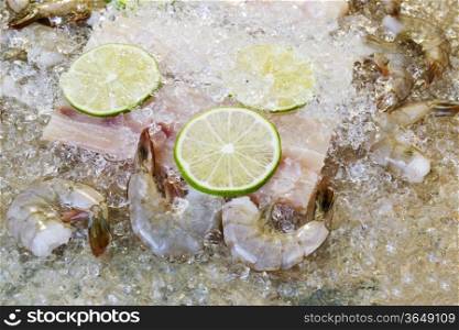 Closeup horizontal photo of fresh raw white fish, shrimp, lime slices and crushed ices on top with natural stone underneath as background