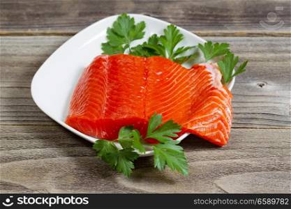Closeup horizontal photo of fresh red salmon fillet on white plate with parsley on the side and rustic wood underneath