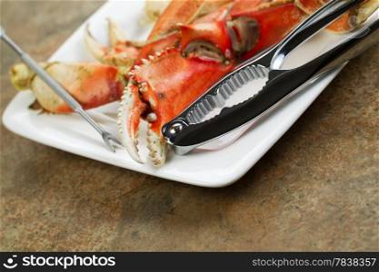 Closeup horizontal photo of freshly cooked Dungeness crab legs on white plate with stainless crab crackers and stone counter top underneath