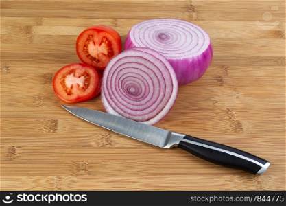 Closeup horizontal photo of freshly sliced red onion and tomato with kitchen knife in front and natural bamboo wood underneath
