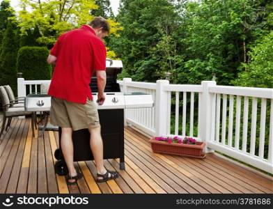 Closeup horizontal photo of mature man working on BBQ grill on open cedar patio with seasonal trees in full bloom in background