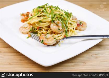 Closeup horizontal photo of Pasta dish with focus on large shrimp in stainless steel fork, basil, and parsley inside of white square plate with natural bamboo wood in background
