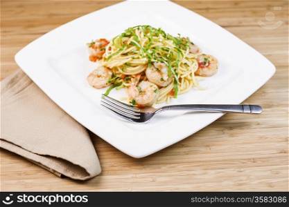 Closeup horizontal photo of Pasta dish with large shrimp, basil, parsley, stainless steel fork and cloth napkin next to white square plate with natural bamboo wood in background