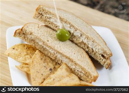 Closeup horizontal photo of tuna fish sandwich, focus on single green olive, and chips, with natural bamboo cutting board underneath and kitchen counter top in background