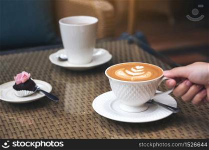 Closeup image of a hand holding a cup of hot latte coffee with latte art and snack on table in cafe