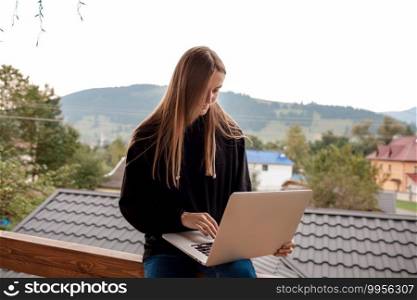 Closeup image of a woman working and typing on laptop while sitting on wooden balcony with green mountains on foggy day with blue sky.. Closeup image of a woman working and typing on laptop while sitting on wooden balcony with green mountains on foggy day with blue sky