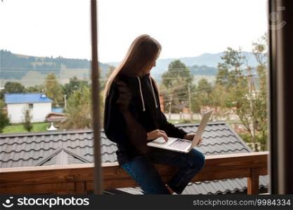 Closeup image of a woman working and typing on laptop while sitting on wooden balcony with green mountains on foggy day with blue sky.. Closeup image of a woman working and typing on laptop while sitting on wooden balcony with green mountains on foggy day with blue sky