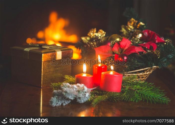 Closeup image of burning candles, Christmas wreath and golden gift box on table next to burning fireplace at living room. Image with shallow depth of focus.