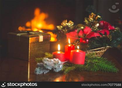Closeup image of burning candles, Christmas wreath and golden gift box on table next to burning fireplace at living room. Image with shallow depth of focus.