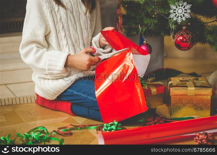 Closeup image of girl sitting under Christmas tree and cutting red paper with scissors for decorating presents