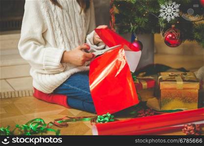 Closeup image of girl sitting under Christmas tree and cutting red paper with scissors for decorating presents