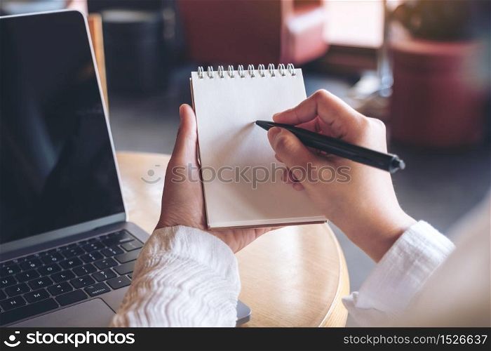 Closeup image of hands holding and writing down on a blank notebook with computer laptop on table