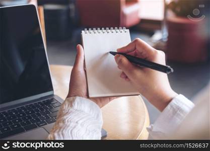 Closeup image of hands holding and writing down on a blank notebook with computer laptop on table
