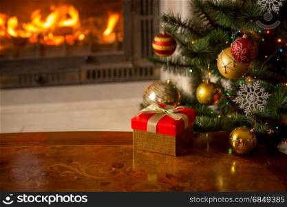Closeup image of red gift box on wooden table in front of burning fireplace and Christmas tree. Empty place for text