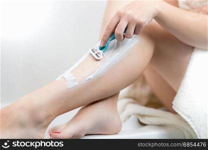 Closeup image of young woman sitting on bath side and shaving legs