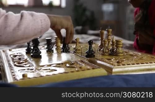 Closeup little girl playing chess making a move holding black rook in her hand. Chess pieces on the board. African american child&acute;s hand moving black rook two fields forward in chess game while playing with her sister at home. Dolly shot. Slo mo.