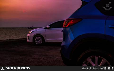 Closeup luxury blue SUV and white car parked on concrete parking area beside the beach in the evening with beautiful purple sunset sky . Road trip travel on summer vacation at tropical sea beach.