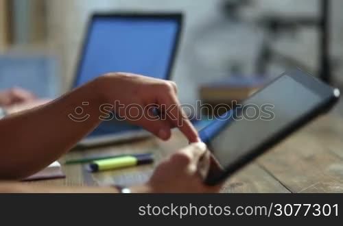 Closeup male student&acute;s hands using touchpad over blurred background with a friend working on laptop. Male freelancer working on digital tablet at home interior. Young teenage hipster scrolling and zooming on touchscreen device.