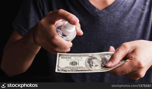 Closeup man wearing protective face mask applying sanitizer spray to 100 dollar money for cleaning, hygiene prevention COVID-19 virus or coronavirus protection concept, dark on black background