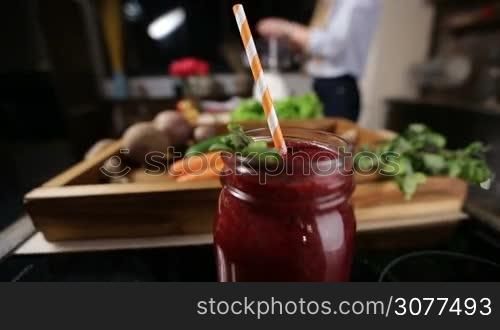 Closeup mason jar of healthy fresh beetroot smothie with striped raw and mint leaves for decoration over wooden tray with food ingredients and blurry woman cooking in the kitchen background. Organic food and healthy eating lifestyle.
