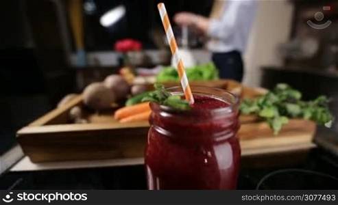 Closeup mason jar of healthy fresh beetroot smothie with striped raw and mint leaves for decoration over wooden tray with food ingredients and blurry woman cooking in the kitchen background. Organic food and healthy eating lifestyle.