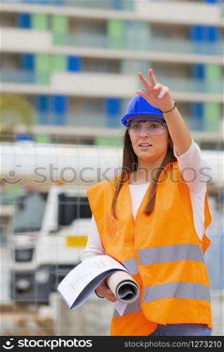 Closeup of a beautiful teenage architecture student holding blueprints and wearing safety helmet, glasses and vest raising her arm on an out of focus background. Work and apprenticeship concept.. Closeup of an architecture student with a raised arm