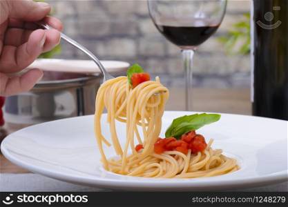 Closeup of a hand picking up spaghetti from a dish in with an out of focus glass of wine, a bottle and a pot in the background. Vegetarian and culinary concept.. Using a fork to pick up spaghetti from a dish