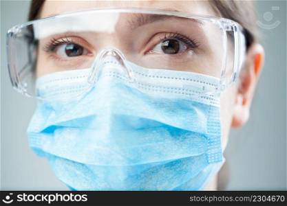 Closeup of a worried female NHS UK doctor or lab scientist wearing PPE safety glasses   blue face mask,COVID-19 corona virus disease global pandemic outbreak,uncertainty anxiety   lost hope concept