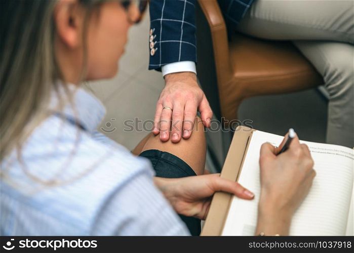 Closeup of boss putting hand over legs of blonde secretary in the office. Sexual harassment at work concept.. Boss sexual harassing to blonde secretary at workplace