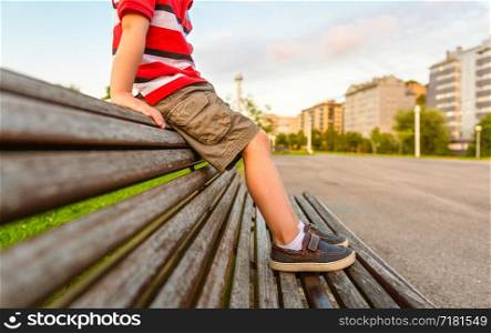 Closeup of boy legs with short pants sitting on the top of wooden bench park relaxing in a bored summer day. Boy legs sitting on the top of bench park relaxing