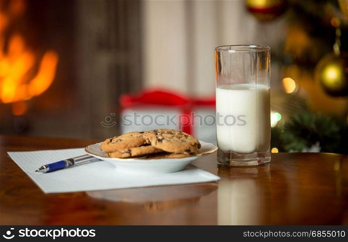 Closeup of cookies, glass of milk and letter for Santa Claus on table next to burning fireplace and Christmas tree