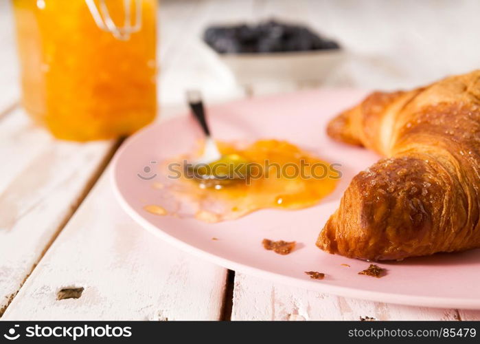 Closeup of croissant and orange jam on a pink plate over a wooden table. Closeup of croissant and jam on a pink plate